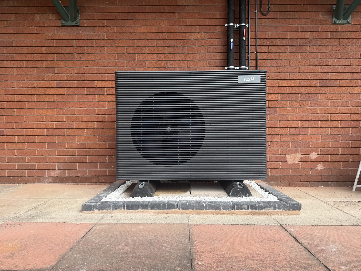 A neatly installed dark heat pump centred against a brick wall